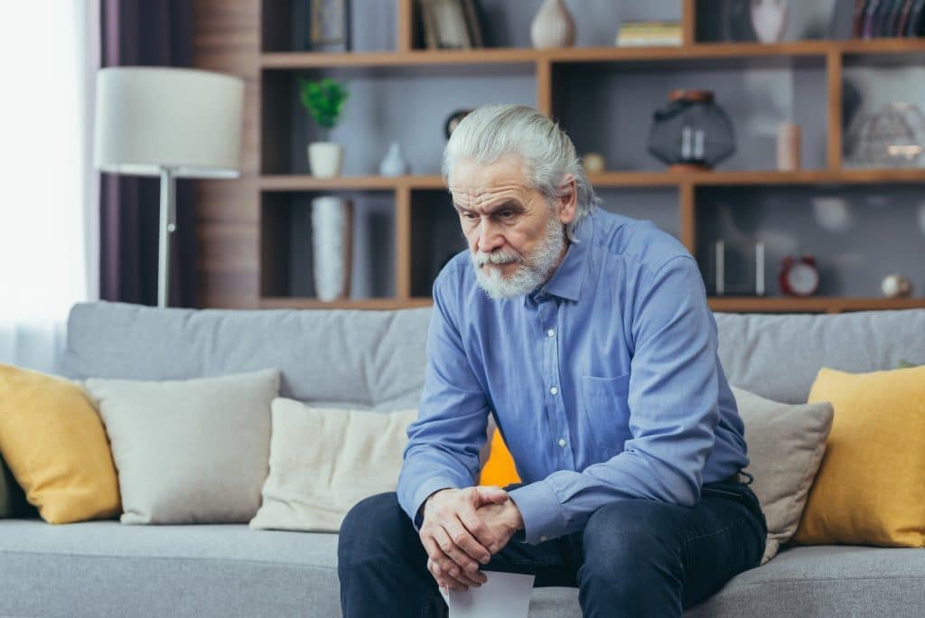 Older man sitting alone on a grey couch with pillows staring out in front of him