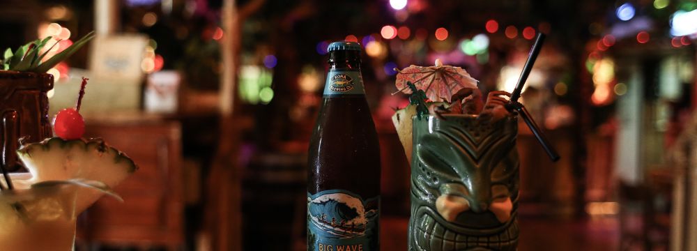 Drinks sitting at the bar top of a tiki bar, one in a bottle, one in a tiki glass